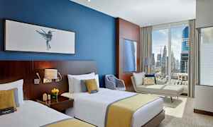 Et hotellrom med to enkeltsenger, en blå vegg med bilder, en sofa, en lenestol og et stort vindu med utsikt over en by. (AI-generert tekst)