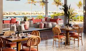 En uteservering med bord og stoler, sofaer og utsikt over vann og hus. (AI-generert tekst)