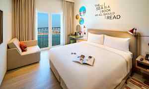 Et hotellrom med en stor seng, sitteplass og åpent vindu med utsikt over havet. (AI-generert tekst)