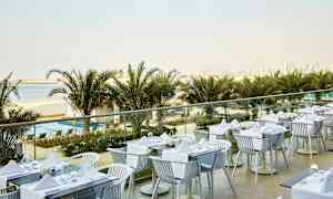 En utendørs restaurant med dekkede bord, med utsikt over palmer, en strand og en bysilhuett i bakgrunnen. (AI-generert tekst)