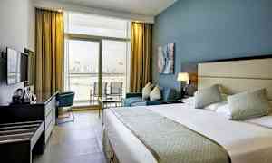 Et hotellrom med en stor seng, en sittegruppe og en arbeidsplass med en vannkoker. Rommet har et stort vindu som vender mot utsikten over en by ved vannet. (AI-generert tekst)