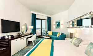 Et moderne hotellrom med en stor seng, TV, arbeidsområde og en sittegruppe ved et vindu som åpner mot en balkong med havutsikt. (AI-generert tekst)