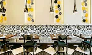 En restaurantinteriør med svart-hvite gulvfliser, mønstrede veggpaneler og en dekorativ sittebenk med svarte stoler rundt bordene. Fargerike veggdekorasjoner vises på veggen. (AI-generert tekst)