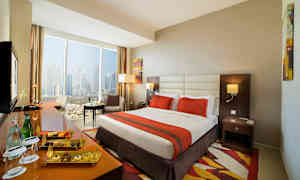 Hotellrom med stor seng, oransje sengeteppe, vindu med utsikt over byen og sittegruppe. (AI-generert tekst)