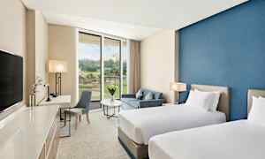 Et hotellrom med to enkeltsenger, en blå vegg, sittegruppe med bord og sofa samt et stort vindu som viser utsikten. (AI-generert tekst)