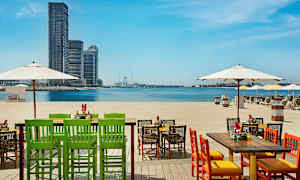 Stoler og bord på en strand med utsikt over havet og byens silhuett i det fjerne. (AI-generert tekst)