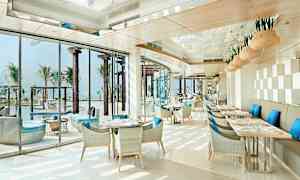 En lys og moderne restaurant med blå puter på stolene og utsikt over et basseng og palmer. (AI-generert tekst)