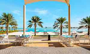 Uteservering med bord, stoler og utsikt over tropiske palmer, hvit sandstrand og hav. (AI-generert tekst)