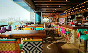 En moderne restaurant med fargerike stoler og barstoler, tremøbler og utsikt over en bysilhuett ved vannet. (AI-generert tekst)