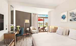 Et hotellrom med moderne møbler, stor seng, marmorbord og en terrasse med solsenger og utsikt over palmer. (AI-generert tekst)