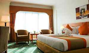 Et hotellrom med en stor seng, to lenestoler, et rundt bord og et stort vindu med gardiner. (AI-generert tekst)