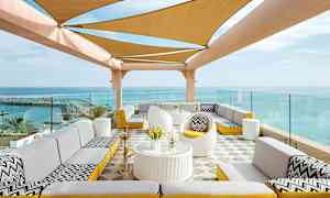 En uteplass med sittegrupper og en skyggetak dekket med stoff foran en havutsikt. (AI-generert tekst)