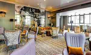 Et moderne hotellrom med komfortable sittemøbler, elegante dekorasjoner og en veggtapet med tekst og kunst. Store vinduer slipper inn naturlig lys. (AI-generert tekst)