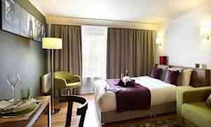 Et hotellrom med en dobbeltseng med lilla og hvite sengetøy, en grønn stol og et spisebord med servise. (AI-generert tekst)