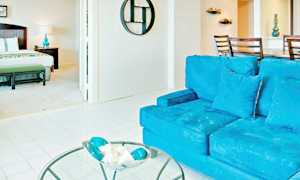 En lys og romslig interiør med en stue og et soverom. I forgrunnen ses en blå sofa samt et glassbord med dekorasjoner. Soverommet har en seng og en nattlampe. (AI-generert tekst)