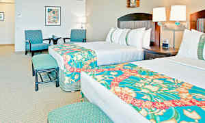 Hotellrom med to senger, dekorert med fargerike sengetepper. Rommet har også en sittegruppe med to stoler og et bord samt veggdekorasjoner. (AI-generert tekst)