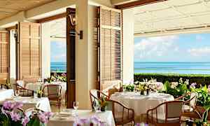En restaurant med bord dekket med hvite duker og blomster. Bakgrunnen viser havutsikt med blå himmel. (AI-generert tekst)