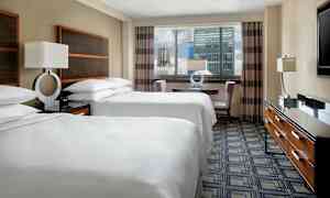 Et hotellrom med to dobbeltsenger kledd i hvitt sengetøy, et bord med stoler nær et vindu og en kommode med en TV på toppen. (AI-generert tekst)