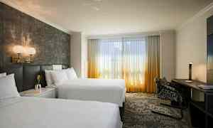 Hotellrom med to store senger, skrivebord og store vinduer med gardiner som slipper inn dagslys. (AI-generert tekst)