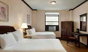 Et hotellrom med to dobbeltsenger, et skrivebord med en stol, et skap og en vegglampe. (AI-generert tekst)