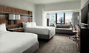 Et hotellrom med to dobbeltsenger, en sittegruppe, et skrivebord og en TV, med utsikt over en by gjennom vinduet. (AI-generert tekst)