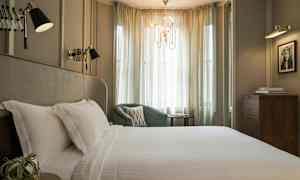 Et elegant soverom med en stor seng dekket med hvite laken, en grønn lenestol og myke gardiner som slipper inn lys. (AI-generert tekst)