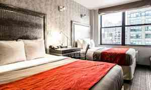 Et hotellrom med to senger med hvitt sengetøy og oransje sengetepper. Det er et vindu med utsikt over en by. (AI-generert tekst)