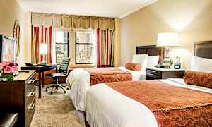 Hotellrom med to enkeltsenger, et skrivebord med stol, samt en lampe og vinduer mot byen. (AI-generert tekst)