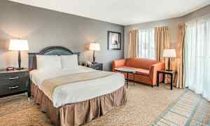 Et hotellrom med en dobbeltseng, et nattbord på hver side, en liten sofa, salongbord og tre lamper. Vinduer med gardiner er synlige i bakgrunnen. (AI-generert tekst)