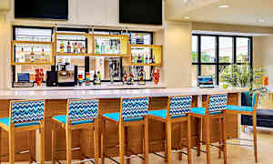 En bar med høye stoler i tre og blå ryggstøtter, en benkeplate med diverse drikkevarer og en kaffemaskin. (AI-generert tekst)