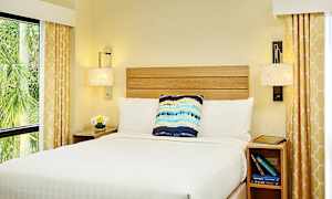 Et soverom med en seng oppredd med hvite sengetepper, en dekorativ pute og nattbord. Gardiner med gult mønster rammer inn vinduene. (AI-generert tekst)