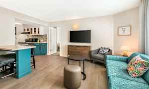 Stue med en sofa, lenestol og en TV på veggen. Et kjøkkenområde med blå skap vises til venstre. (AI-generert tekst)