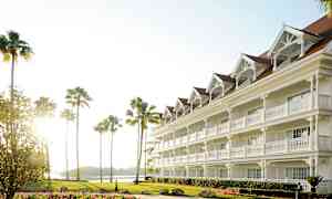 Et hvitt hotell med flere etasjer og innganger med verandaer, omgitt av palmer og en plen med blomster. (AI-generert tekst)