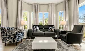 En moderne stue med en sofa og to lenestoler, et salongbord i midten og store vinduer som slipper inn naturlig lys. (AI-generert tekst)