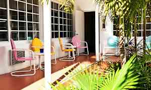 Fargerike metallstoler og et bord står på en terrasse foran et hus med store vinduer. Foran terrassen er grønne palmer. (AI-generert tekst)