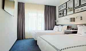 Et hotellrom med to enkeltsenger med hvitt sengetøy og dekorative overkast. Rommet har mørkelilla gardiner, en veggmontert TV og innrammede bilder på veggen over sengegavlen. (AI-generert tekst)