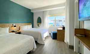 Et moderne hotellrom med to dobbeltsenger, et skrivebord og et stort vindu som slipper inn naturlig lys. (AI-generert tekst)