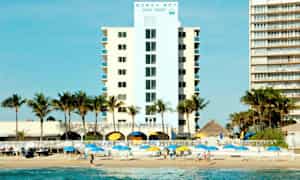 Et hotell med palmer foran, ved siden av stranden med parasoller og mennesker på sanden. (AI-generert tekst)