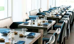 Et elegant restaurantområde med dekkede bord, blå servietter og glass. Lyse vinduer i bakgrunnen. (AI-generert tekst)