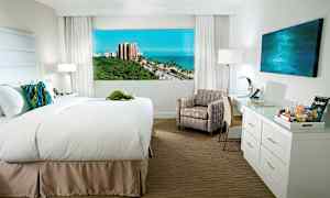 Hotellrom med en bred seng, stol, skrivebord og utsikt over havet og grønne områder utenfor. (AI-generert tekst)