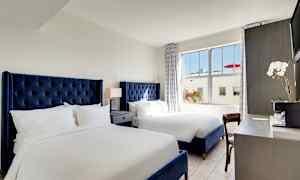 Et hotellrom med to senger med blå hodegjerder, et nattbord med lampe mellom sengene og et skrivebord med en blomst og kaffetrakter. (AI-generert tekst)