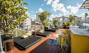 En terrasse med solsenger, puter og grønne planter. Det er en gul bardisk med krakker på siden. (AI-generert tekst)