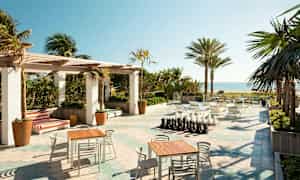 En utendørsterrasse med bord og stoler, et sjakkbrett og utsikt over stranden og havet, omgitt av palmer. (AI-generert tekst)