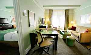 Et hotellrom med lys innredning, inkludert en seng, skrivebord, sofa og TV. (AI-generert tekst)