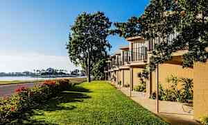 Bildet viser et grønt område med plen, en rekke bygninger med balkonger og en vei som går ved siden av. I bakgrunnen sees vannet og palmer. (AI-generert tekst)