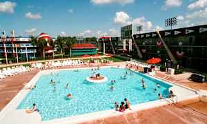 Utendørsbasseng med solsenger rundt og folk som svømmer. I bakgrunnen sees bygninger med sportstema og en stor dekorativ gjenstand som ligner en amerikansk fotball samt palmer. (AI-generert tekst)