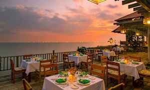 En uteservering med dekkede bord på en treterrasse ved havet under en fargerik solnedgang. (AI-generert tekst)