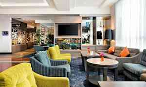 Et hotellobby med komfortable sitteplasser, flere sofaer og stoler i ulike farger, samt bord med blomster. En TV er montert på veggen og det finnes dekorative gjenstander på hyllene. (AI-generert tekst)