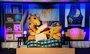 To tegneseriefigurer, en giraff og en bjørn, sitter i en seng med dyne, og scenerekvisitter er synlige i bakgrunnen. (AI-generert tekst)