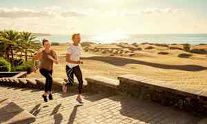 To personer jogger på en steinlagt gangvei nær sanddyner og palmer ved kysten. (AI-generert tekst)